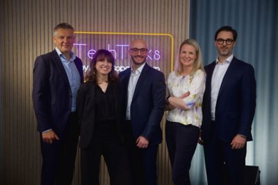 The host and guests of Tech Talks episode 1 posing for a group photo in the studio, in front of a neon style sign saying "TechTalks". The group consists of three men and two women, wearing office apparel and smiling.