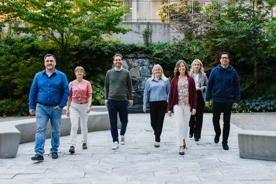 A group of happy employees are walking towards the camera in an outdoor area with trees, bushes and a fountain. ​