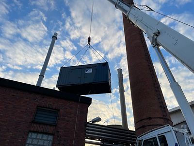 Frog perspective of a hot water boiler being lifted by a large crane at an industrial site. The sky is clear blue.