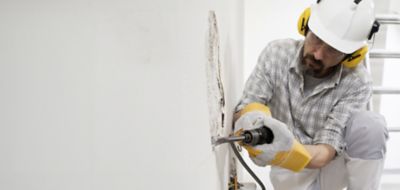 house renovation concept, construction worker breaks the old plaster of the wall with pneumatic air hammer chisel, wears gloves, helmet and safety yellow headphones