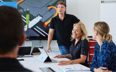 Employees seated in a meeting room with one employee standing by the screen