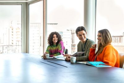 Drei junge Auszubildende sitzen gemeinsam im modernen Büro, lachen und arbeiten an Unterlagen​.