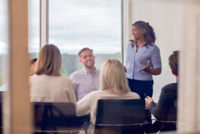 A woman employee stands and speaks while others listen around a meeting table in a glass-walled room.