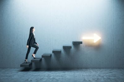 Young businesswoman moving up on stairs with arrow on gray wall. Leadership and career development concept.