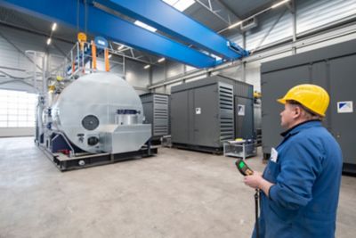 Operator wearing a yellow helmet monitoring an industrial steam boiler at the workshop of Eco Steam and Heating with multiple boilers in the background.