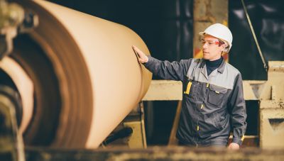A man wearing a white hard hat and work uniform checks a large roll of paper on a machine in a paper mill.