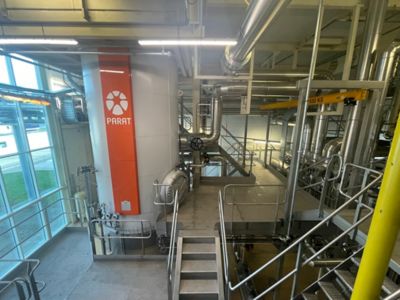 Industrial boiler labeled "PARAT" in a facility with metal walkways and pipes. Large windows allow natural light inside.