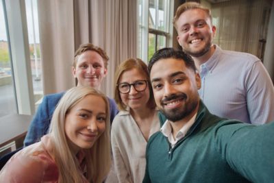 A group of five employees pose for a cheerful selfie near a window inside a workplace setting