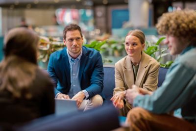 Male and female employees in focus, seated on a sofa in a plant-lined office lounge, with two other colleagues blurred in the foreground