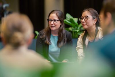 Two female employees in focus, seated in an indoor workspace with greenery in the background, surrounded by blurred colleagues in front