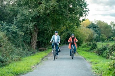 Two people in helmets cycling down a road with greenery on both sides.