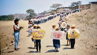 Dirt road lined with people observing children with smiles on their faces walking down the road towards the camera holding handwritten messages. 