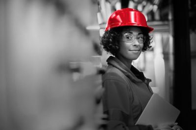 A woman in a red hardhat is leaning against a wall and smiling at the camera.