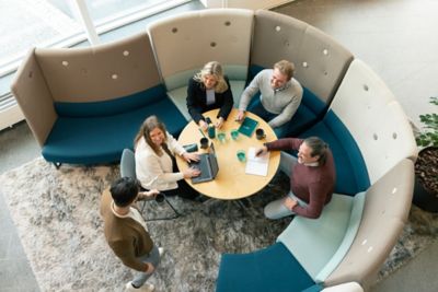 Four colleagues sitting around a round table working together, as seen from above. One person is working on a laptop, the others have notebooks. They look up to greet a fifth colleague, who is about to join them.  ​