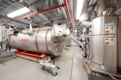 Two large Bosch boilers in a boiler room. The boilers are made of stainless steel and have a variety of pipes and valves attached to them.