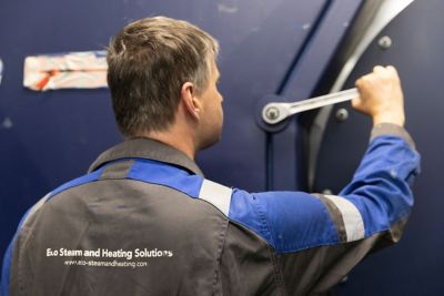 Technician from Eco Steam and Heating Solutions adjusts equipment with a wrench. The company logo is visible on the back of his uniform.