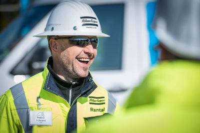 Close-up of a service technician wearing a white hard hat. Atlas Copco Rental logo is visible on the safety jacket; another worker is partially visible.