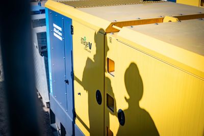 Gros plan d’un générateur jaune « conforme aux normes Stage V » à côté d’une machine bleue, avec deux ombres de casques de sécurité projetées sur le générateur.