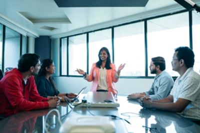 Team meeting in conference room with woman standing at the center seemingly speaking. 