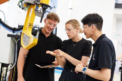 Three employees gathered around to look at something one of them is holding in their hand, while standing next to a machine.​