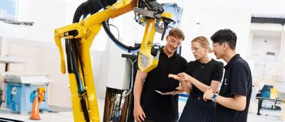 Three employees gathered around to look at something one of them is holding in their hand, while standing next to a machine.​