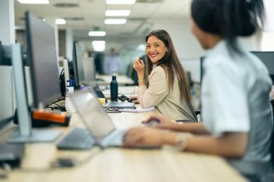 Two women colleagues seated next to each in office smiling