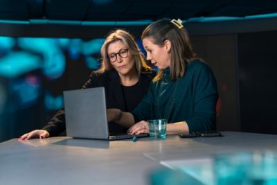 Two female colleagues are looking at a laptop together. Their faces express focus as one woman seems to point at something on the screen while explaining something to the other woman. ​