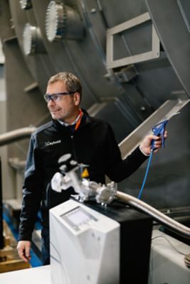 An employee pauses from working on a machine with a handheld tool, to look at someone or something over his shoulder out of frame.​