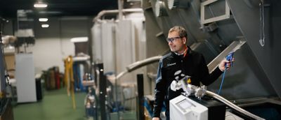 An employee pauses from working on a machine with a handheld tool, to look at someone or something over his shoulder out of frame.​