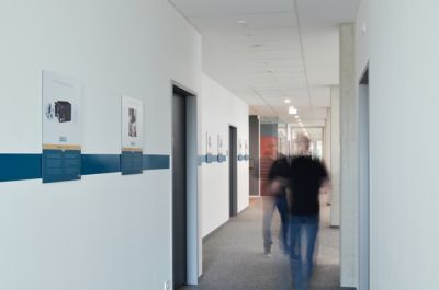 Flur mit weißen Wänden, blauer Streifen und mehreren Türöffnungen, zwei verschwommene Personen gehen darauf zu