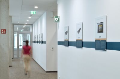 Flur mit weißen Wänden und blauer Streifenlinie, an der Wand hängen mehrere Informationsposter, eine Person in rotem Oberteil und heller Hose geht den Flur entlang
