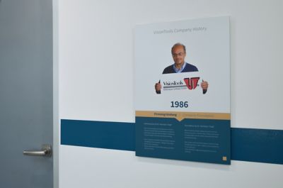 Poster mit einem Mann, der ein Schild mit den Logos von VisionTools hält, an einer weißen Wand in einem Büro mit blauem Streifen und Stühlen im Hintergrund