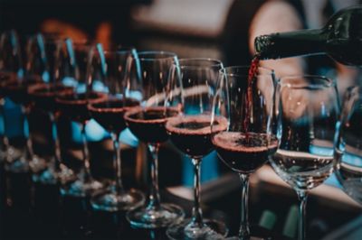 Glasses of red wine lined up in a bar top, with a bottle filling a glass