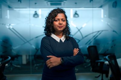 Woman with black curly hair is standing in a conference room with her arms crossed, making eye contact with the camera.​