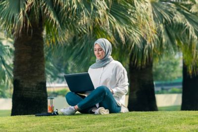 A woman is sitting on the grass in a park working on her laptop.​